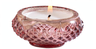 A pink glass candle holder with a textured, diamond-cut pattern and a single lit wick, containing white wax, isolated on a white background.